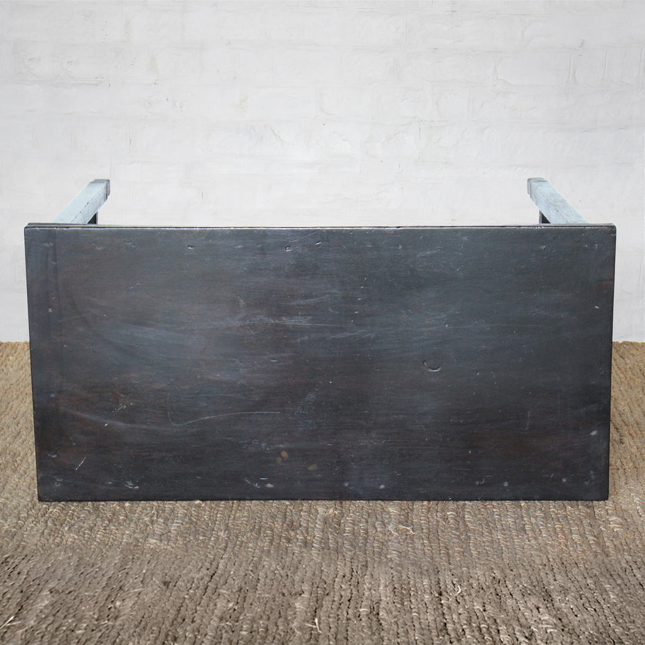 The Chhaya Teak Table by John Robshaw, a handcrafted rectangular black wooden piece inspired by vintage design, is shown from the side, positioned on a textured brown floor with a white brick wall in the background.