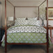 A wooden four-poster bed with patterned green and white bedding features a soft Jeeva Sage Quilt by John Robshaw, adorned with a delicate spring meadow design. Decorative pillows complement the scene, while a bedside table with a lamp on the left and a vase with branches on the right complete the cozy setup.