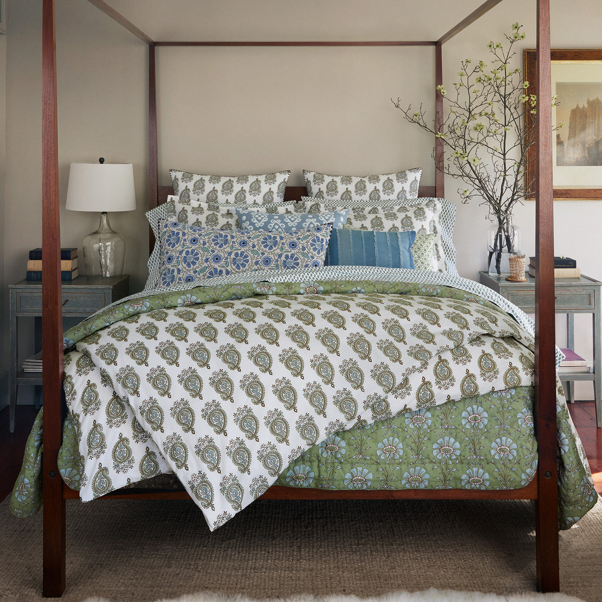 A four-poster bed adorned with the Jeeva Sage Quilt by John Robshaw, showcasing a hand-quilted, spring meadow design in soft cotton, and matching pillows is situated in a room featuring light-colored walls. The ambiance is further enhanced by a table lamp, two nightstands—one of which holds a potted plant—and a framed painting on the wall.