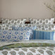 A neatly arranged bed with multiple pillows in various patterns and the John Robshaw Jeeva Sage Quilt, soft cotton featuring floral designs in blue, green, and white hues. Branches with small flowers are placed beside the bed.