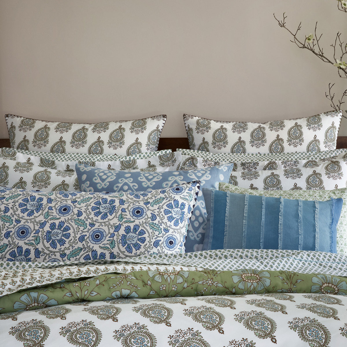 A neatly arranged bed with multiple pillows in various patterns and the John Robshaw Jeeva Sage Quilt, soft cotton featuring floral designs in blue, green, and white hues. Branches with small flowers are placed beside the bed.