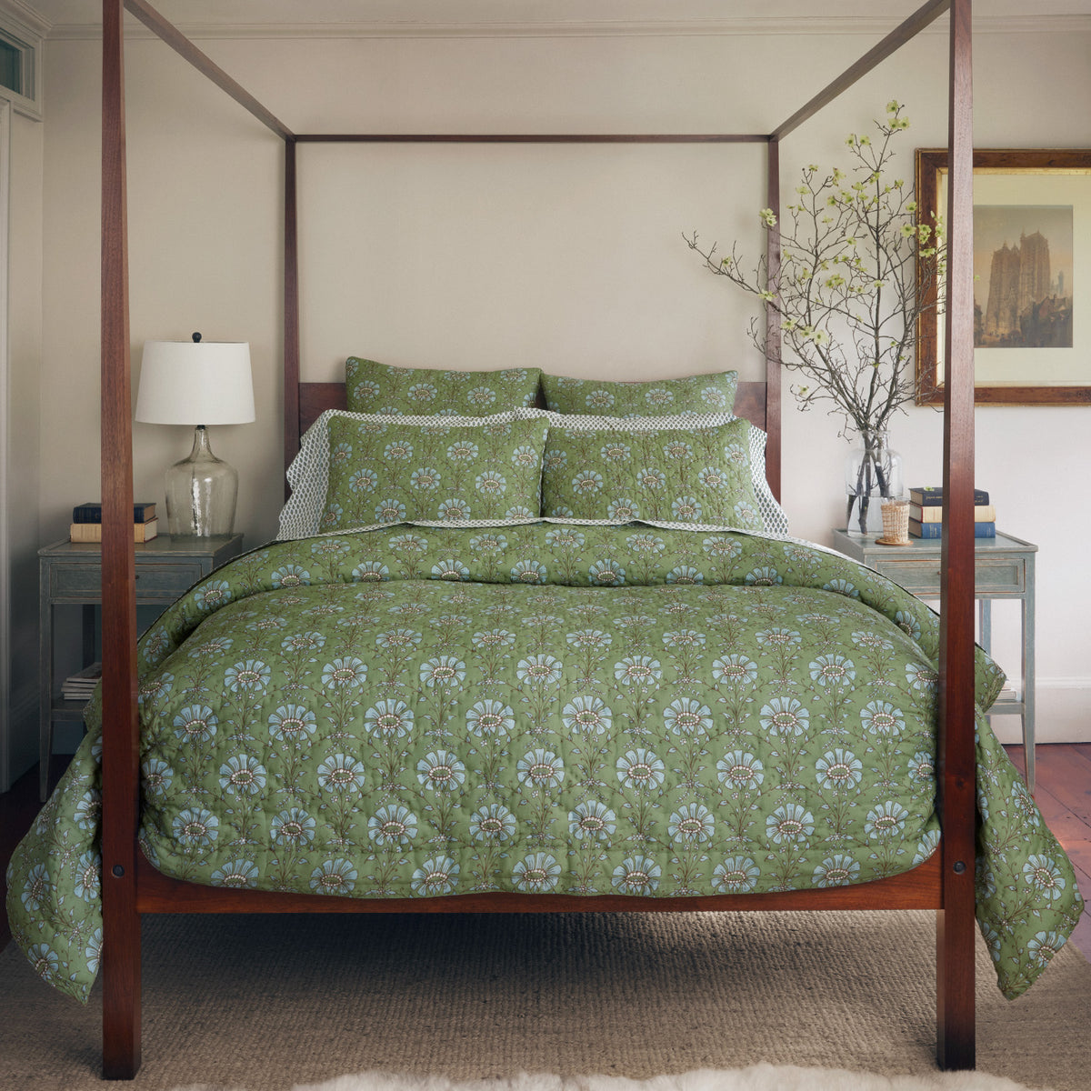 A bedroom with a hand quilted four-poster bed featuring the John Robshaw Jeeva Sage Quilt, a white lamp on a nightstand, a framed picture on the wall, and a branch in a vase on the opposite nightstand.