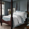 A sophisticated bedroom featuring a wooden four-poster bed, adorned with the John Robshaw Amina Ecru Duvet Set, crafted from the finest quality cotton linen. A nightstand with a lamp, flowers in a vase, and a window with light streaming in complement the light blue walls.