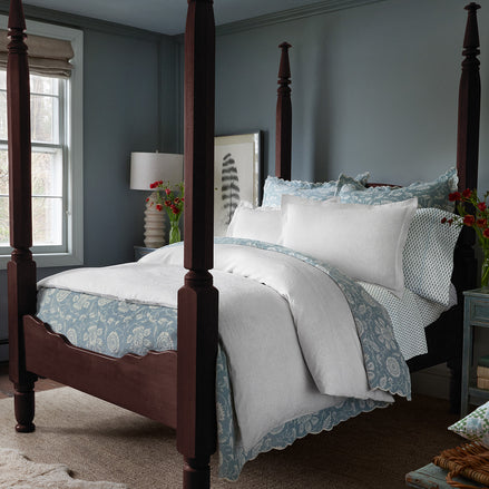 A sophisticated bedroom featuring a wooden four-poster bed, adorned with the John Robshaw Amina Ecru Duvet Set, crafted from the finest quality cotton linen. A nightstand with a lamp, flowers in a vase, and a window with light streaming in complement the light blue walls.