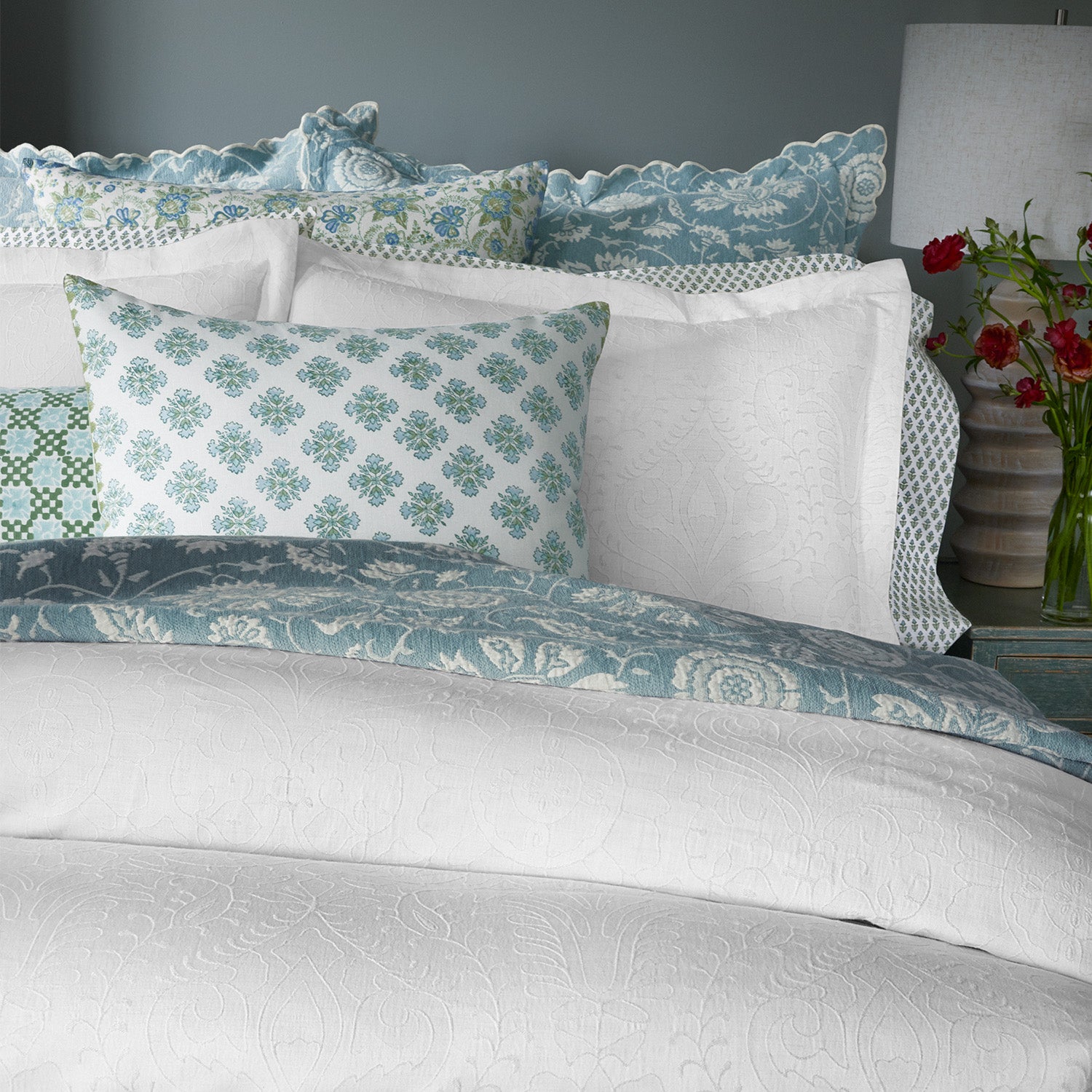 A neatly made bed adorned with multiple pillows in various blue and white patterns, covered by the finest quality cotton linen from the Amina Ecru Duvet Set by John Robshaw, and a lamp with red flowers on a nearby nightstand.