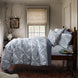 A wooden bed adorned with blue and white patterned bedding, featuring a Dasati Lapis Quilt by John Robshaw, stands next to a bedside table holding books and a glass of water. It is placed near a window with a beige shade, accompanied by a vase filled with branches.
