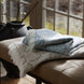 Two folded Dasati Sand throws by John Robshaw, featuring stonewashed cotton with intricate woven designs and fringed edging, are placed on a cushioned bench in front of a window, with a black vase in the background.