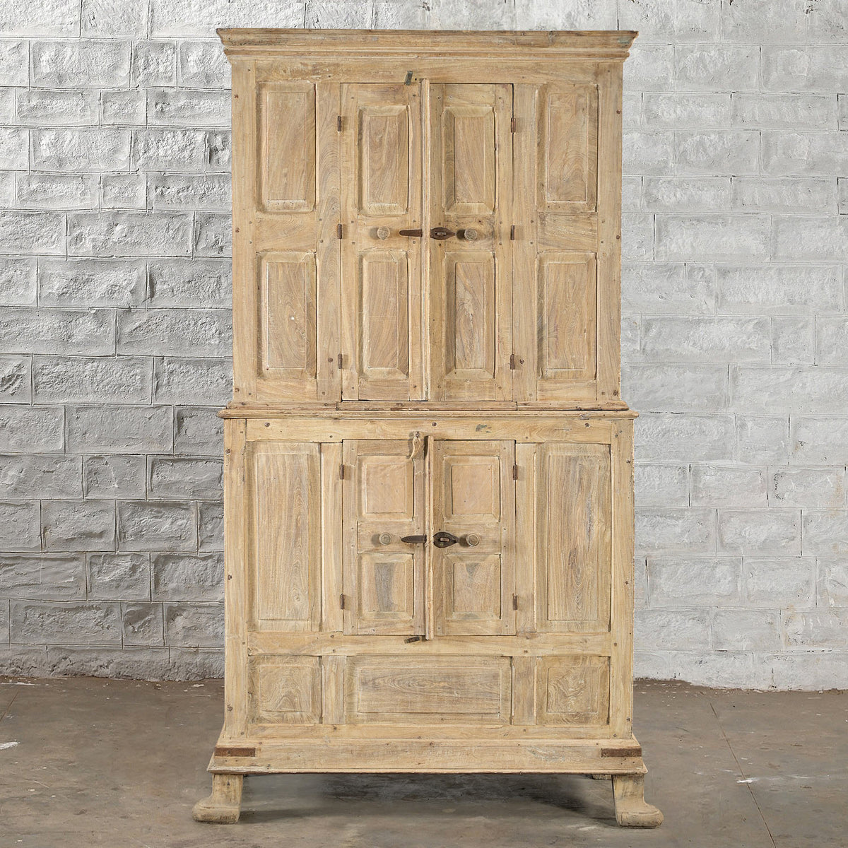 The Capala Teak 2 Part Cabinet by John Robshaw is a tall, rustic piece crafted from bleached teak, featuring paneled doors and metal handles. With its two main sections and refined design, this cabinet stands beautifully against a white brick wall on a concrete floor—a perfect choice for vintage furniture enthusiasts.