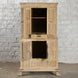 The Capala Teak 2 Part Cabinet by John Robshaw, crafted from tall bleached teak with four doors and a central drawer, is shown open and empty against a white brick wall on a concrete floor. This geometric-style cabinet brings vintage furniture charm to any space.