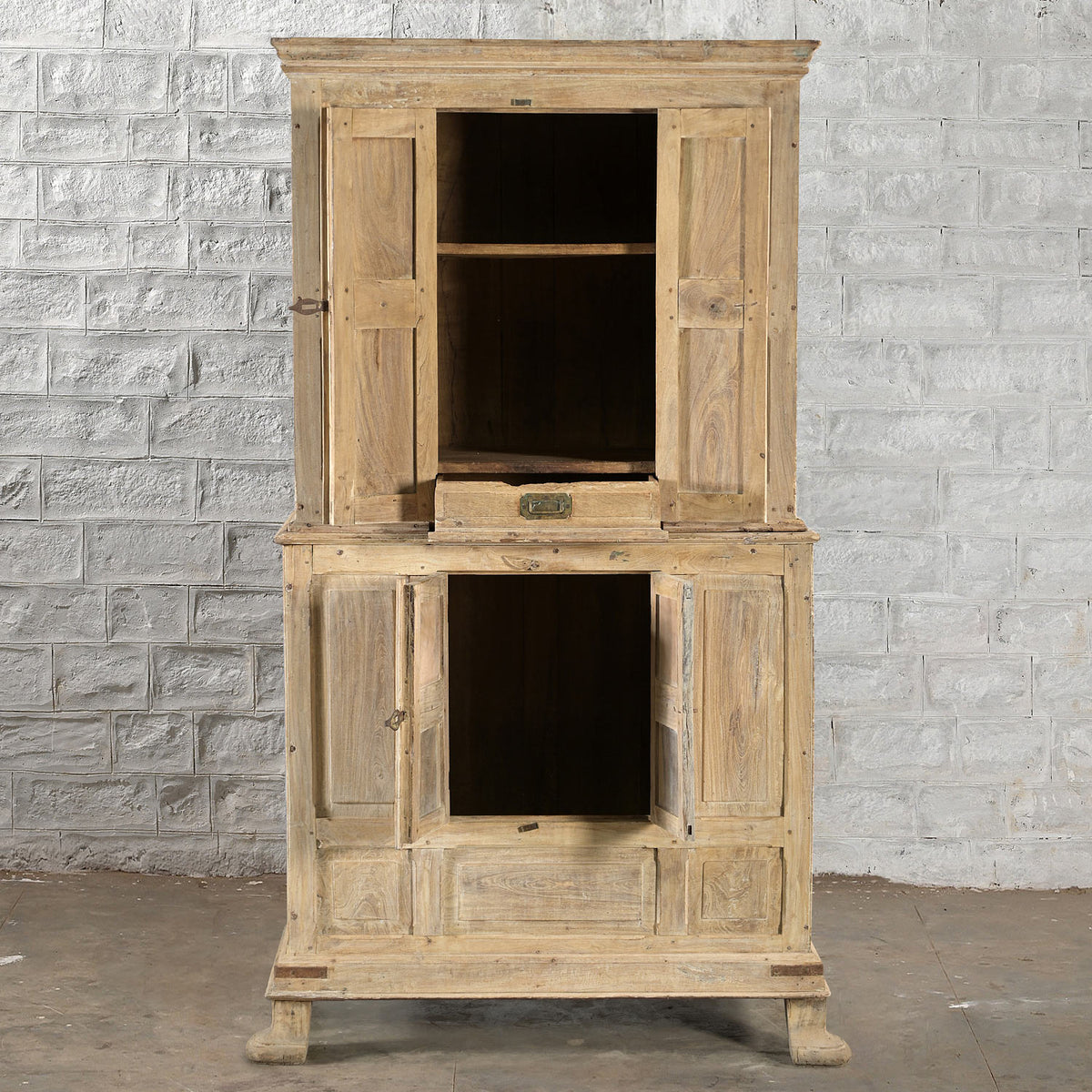The Capala Teak 2 Part Cabinet by John Robshaw, crafted from tall bleached teak with four doors and a central drawer, is shown open and empty against a white brick wall on a concrete floor. This geometric-style cabinet brings vintage furniture charm to any space.