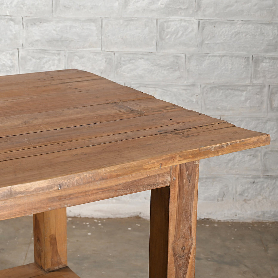 The John Robshaw Teak Bench 2, with its natural, uneven edge, stands on a concrete floor against a gray brick wall, evoking the timeless appeal of handcrafted teak furniture.