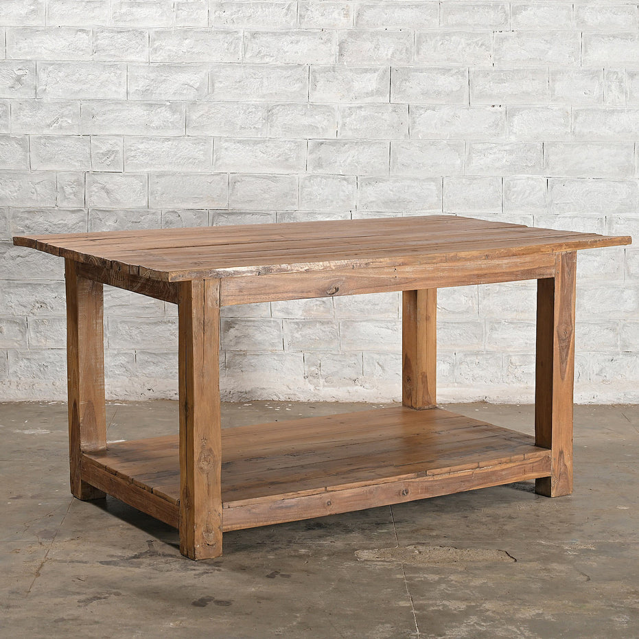 The John Robshaw Teak Bench 2 is a handcrafted teak furniture piece, featuring a lower shelf and standing elegantly on a concrete floor against a white brick wall.