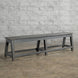 A long, dark gray Madura Teak Bench by John Robshaw, featuring angled legs, sits on a concrete floor against a textured white brick wall, evoking the timeless character of classic school benches.