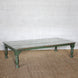 Aangan Teak Coffee Table by John Robshaw, featuring a low rectangular design with weathered green legs and a whitewashed plank-style top, displayed on a woven rug against a white brick wall.