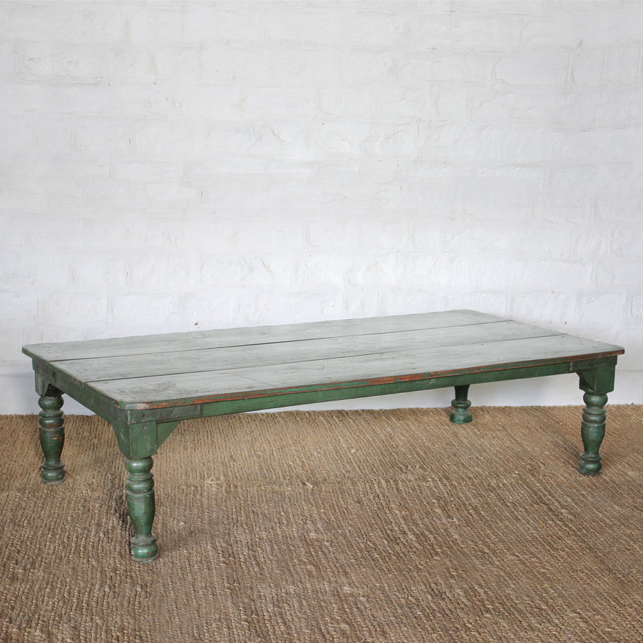 Aangan Teak Coffee Table by John Robshaw, featuring a low rectangular design with weathered green legs and a whitewashed plank-style top, displayed on a woven rug against a white brick wall.