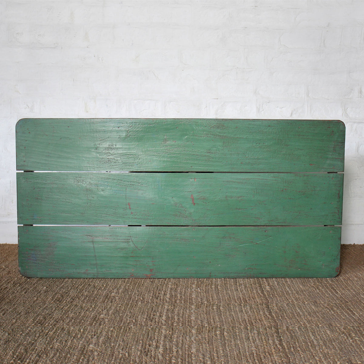 The Aangan Teak Coffee Table by John Robshaw features a plank-style top with three horizontal planks, set against a white brick wall and placed on a brown textured floor.