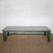 Aangan Teak Coffee Table by John Robshaw sits on a woven rug against a white textured brick wall, featuring a simple silhouette with a weathered green base and a gray plank-style top.