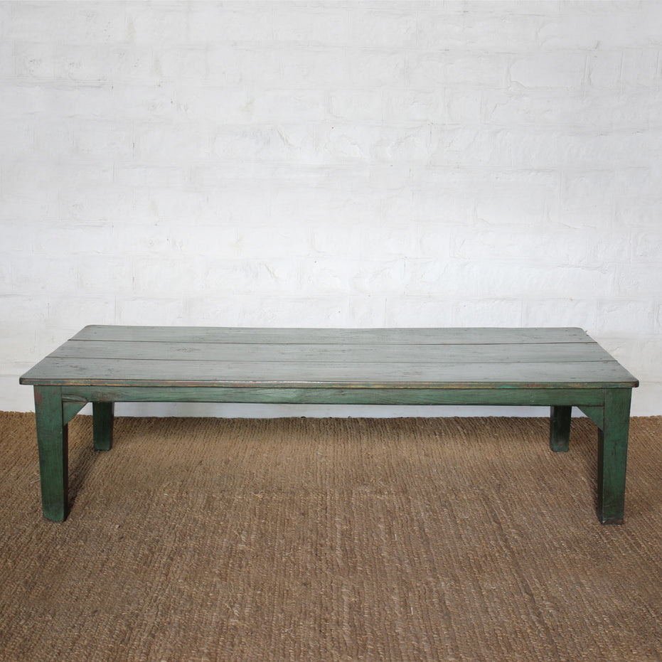 Aangan Teak Coffee Table by John Robshaw sits on a woven rug against a white textured brick wall, featuring a simple silhouette with a weathered green base and a gray plank-style top.