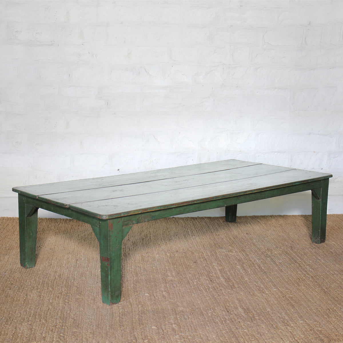 The Aangan Teak Coffee Table by John Robshaw features a low, rectangular design with a weathered green frame and a light-colored, plank-style top, shown placed on a textured brown rug against a white brick wall.