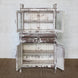 Introducing the Juhi Teak Cabinet by John Robshaw—a character-rich piece crafted in distressed white wood. It features two glass-paneled upper doors, two drawers, and two lower doors, all opening to reveal generous storage and shelving within.
