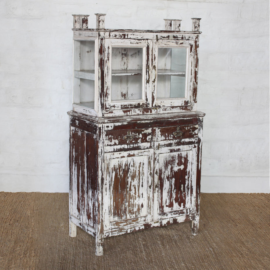 The Juhi Teak Cabinet by John Robshaw is a character-rich piece featuring a distressed wooden finish, glass-paneled doors on top, two drawers, and two solid doors below for ample storage. Standing gracefully on a woven rug against a white brick wall, it brings both charm and functionality to any space.