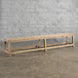 The Umed Teak Bench by John Robshaw, featuring a long, rectangular silhouette and simple design, is handcrafted and sits on a bare concrete floor against a light gray brick wall.