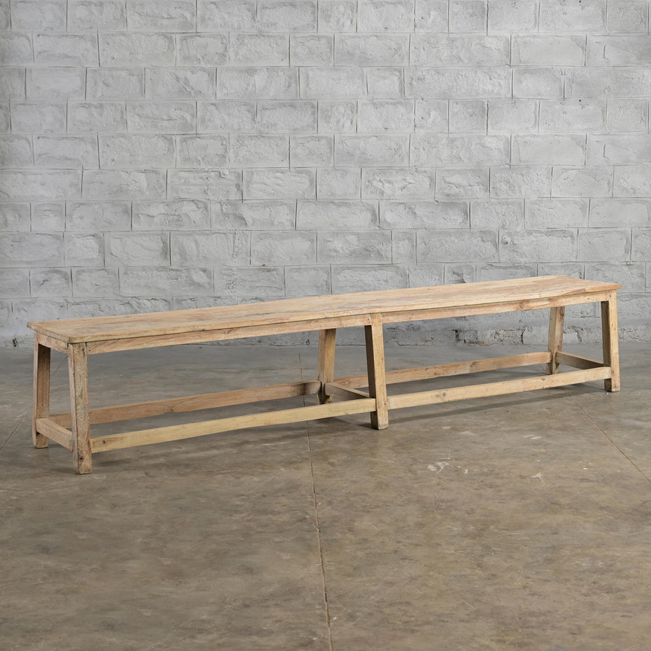 The Umed Teak Bench by John Robshaw, featuring a long, rectangular silhouette and simple design, is handcrafted and sits on a bare concrete floor against a light gray brick wall.