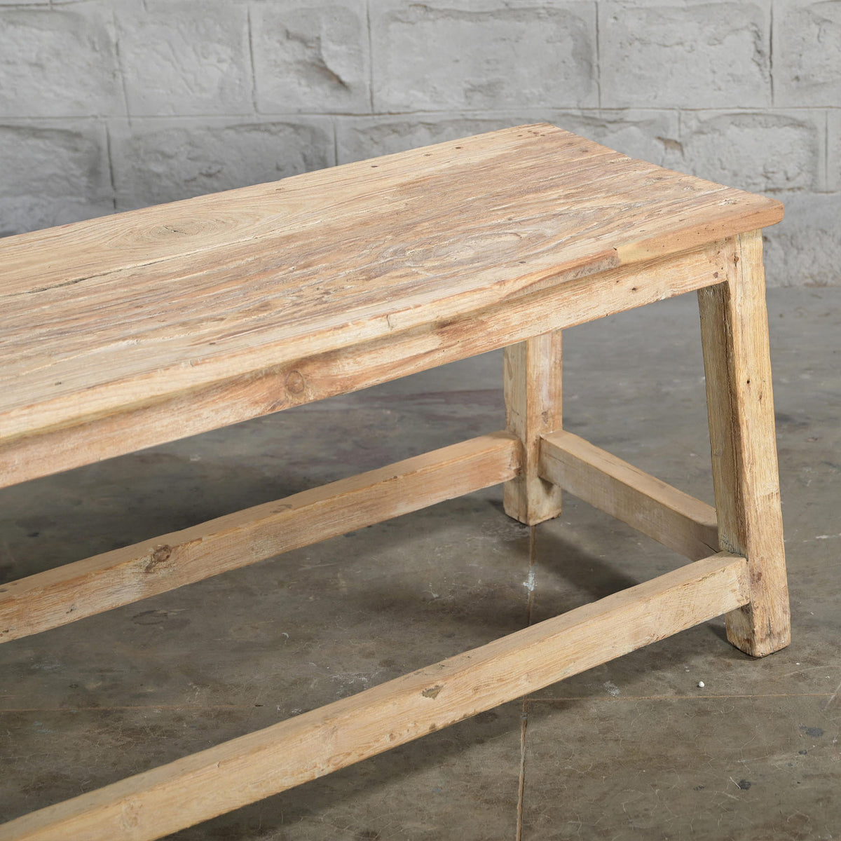 The Umed Teak Bench by John Robshaw, featuring a simple handcrafted design and natural finish, stands on a concrete floor against a textured gray brick wall.