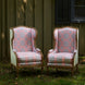 Two Maansi Antique armchairs by John Robshaw, featuring ornate high backs and pink and blue medallion print upholstery, are arranged on grass outside beside a green wooden wall with a shuttered window.