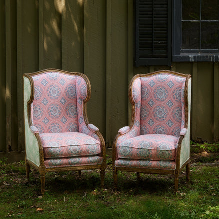Two Maansi Antique and Sage Chairs by John Robshaw, featuring ornate carved wood frames and pink and green patterned antique fabric upholstery, sit side by side on the grass in front of a green wooden wall with a window and black shutter.