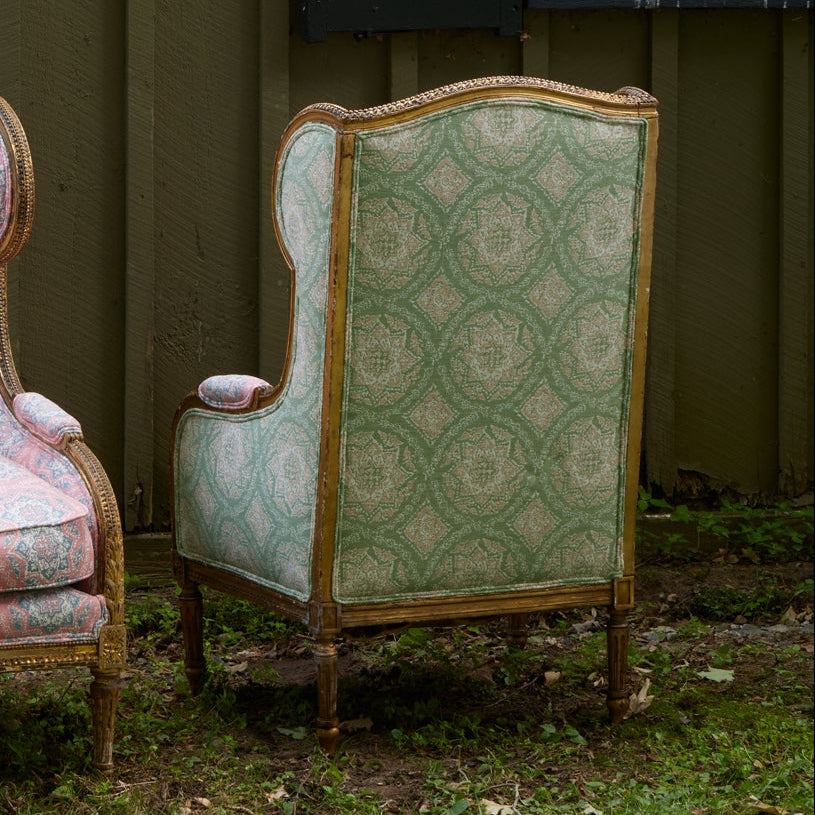 A Maansi Sage armchair by John Robshaw, featuring green block print patterned upholstery, is positioned outdoors on grass with its back facing the camera and a partial view of a matching chair visible to the left.