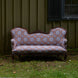 The Maansi Chestnut sofa by John Robshaw, featuring ornate vintage-inspired patterned upholstery and elegant wooden trim, sits on grass outside against a green wooden wall with a black shuttered window—its fabric evocative of classic medallion trade textiles.