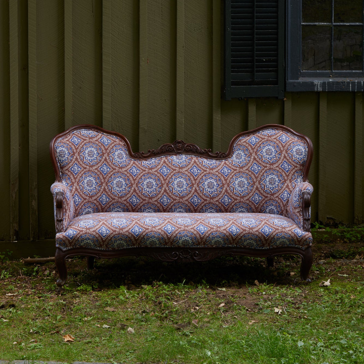 The Maansi Chestnut sofa by John Robshaw, featuring ornate vintage-inspired patterned upholstery and elegant wooden trim, sits on grass outside against a green wooden wall with a black shuttered window—its fabric evocative of classic medallion trade textiles.