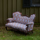 The Maansi Chestnut sofa by John Robshaw, featuring ornate wood trim and blue block print patterned upholstery, sits on grass in front of a green wooden wall and a window.