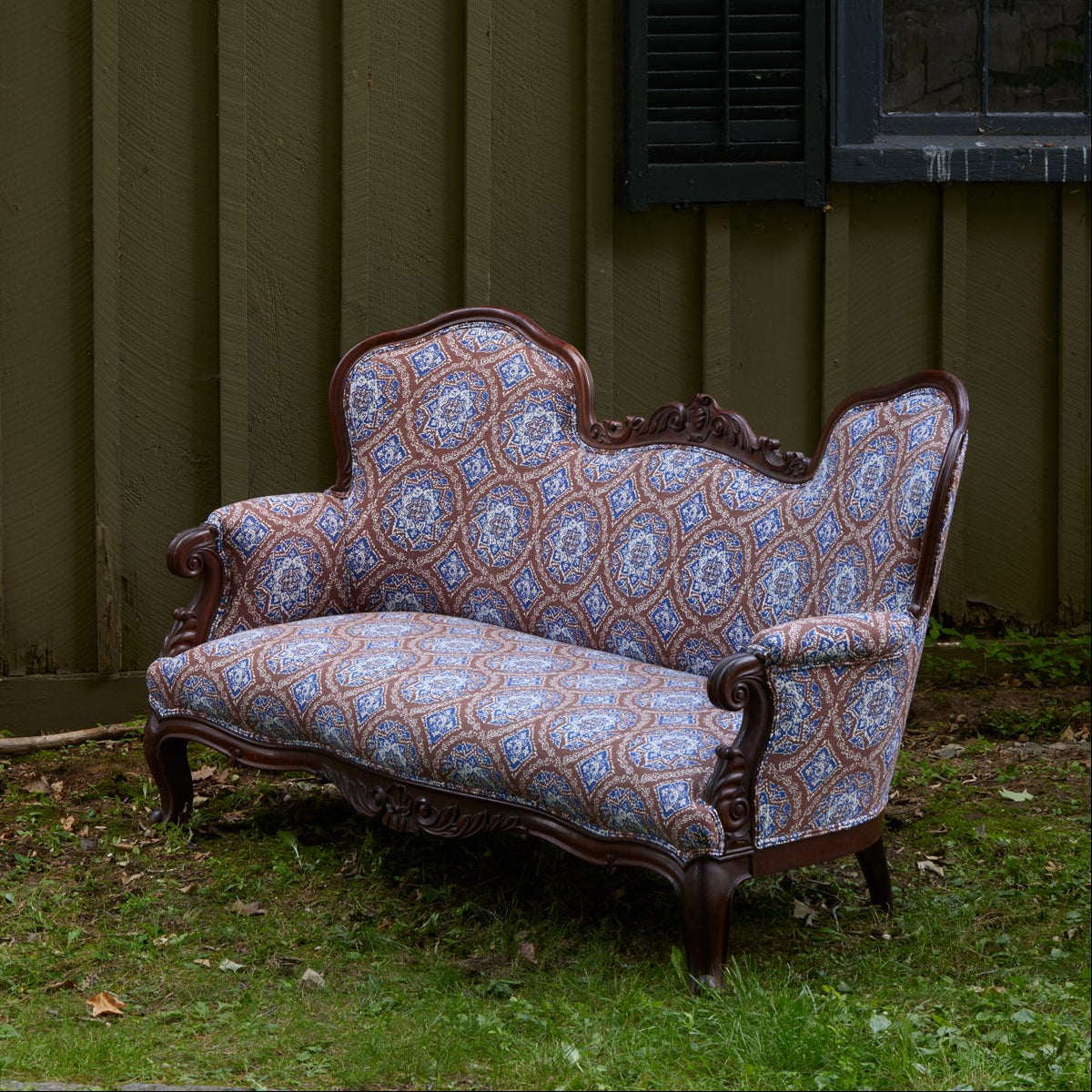 The Maansi Chestnut sofa by John Robshaw, featuring ornate wood trim and blue block print patterned upholstery, sits on grass in front of a green wooden wall and a window.