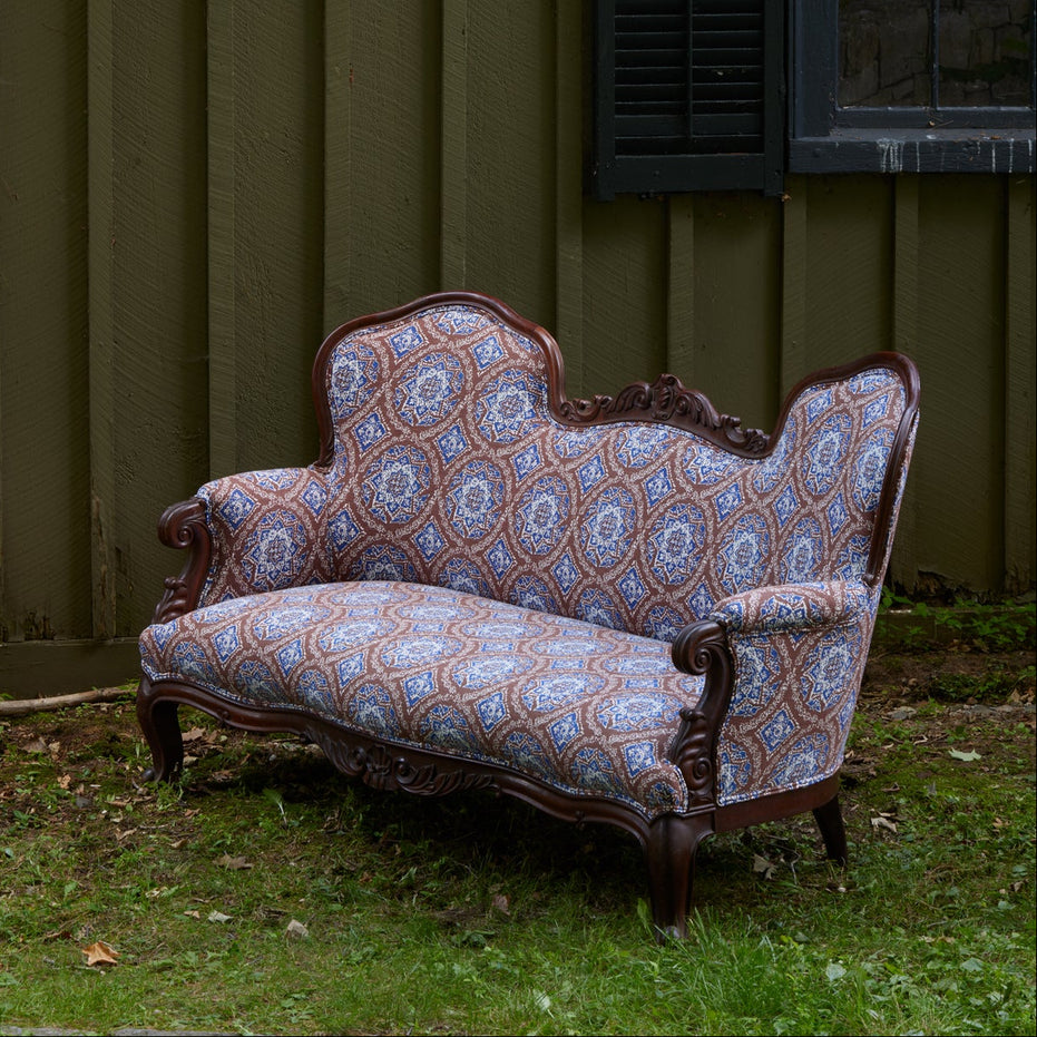 The Maansi Chestnut sofa by John Robshaw, featuring ornate wood trim and blue block print patterned upholstery, sits on grass in front of a green wooden wall and a window.
