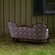 A Maansi Chestnut sofa by John Robshaw, featuring a vintage medallion print pattern, is placed outdoors on grass facing away from the camera, set against a wooden building with a closed shutter and window.
