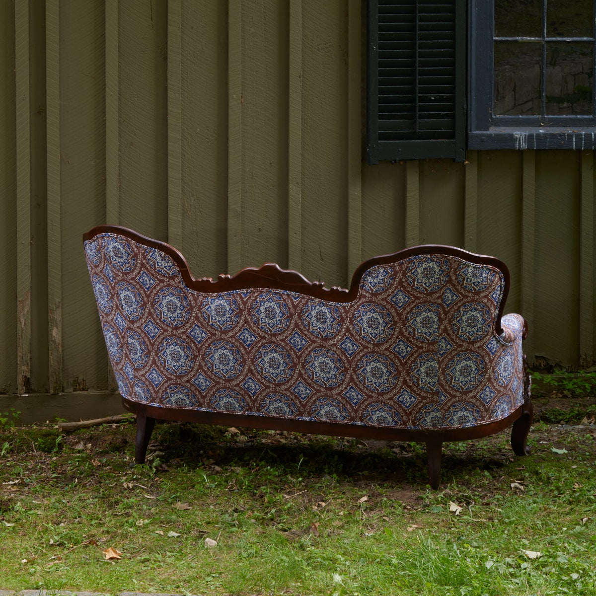 A Maansi Chestnut sofa by John Robshaw, featuring a vintage medallion print pattern, is placed outdoors on grass facing away from the camera, set against a wooden building with a closed shutter and window.