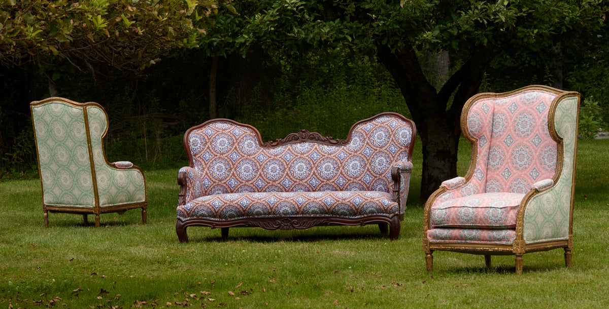A vintage three-piece furniture set consisting of a patterned loveseat in the center and two matching wingback armchairs on either side, displayed outdoors on a green lawn with trees in the background.