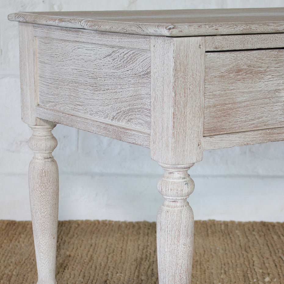 Close-up of the Jharna Teak Desk by John Robshaw—a vintage-inspired writing table crafted from teak with turned legs and a whitewashed finish—situated atop a woven rug against a white brick wall.