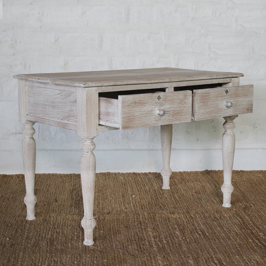 The Jharna Teak Desk by John Robshaw, featuring two open drawers, turned legs, and a distressed finish, sits on a woven rug against a white brick wall, bringing vintage furniture charm to the space.