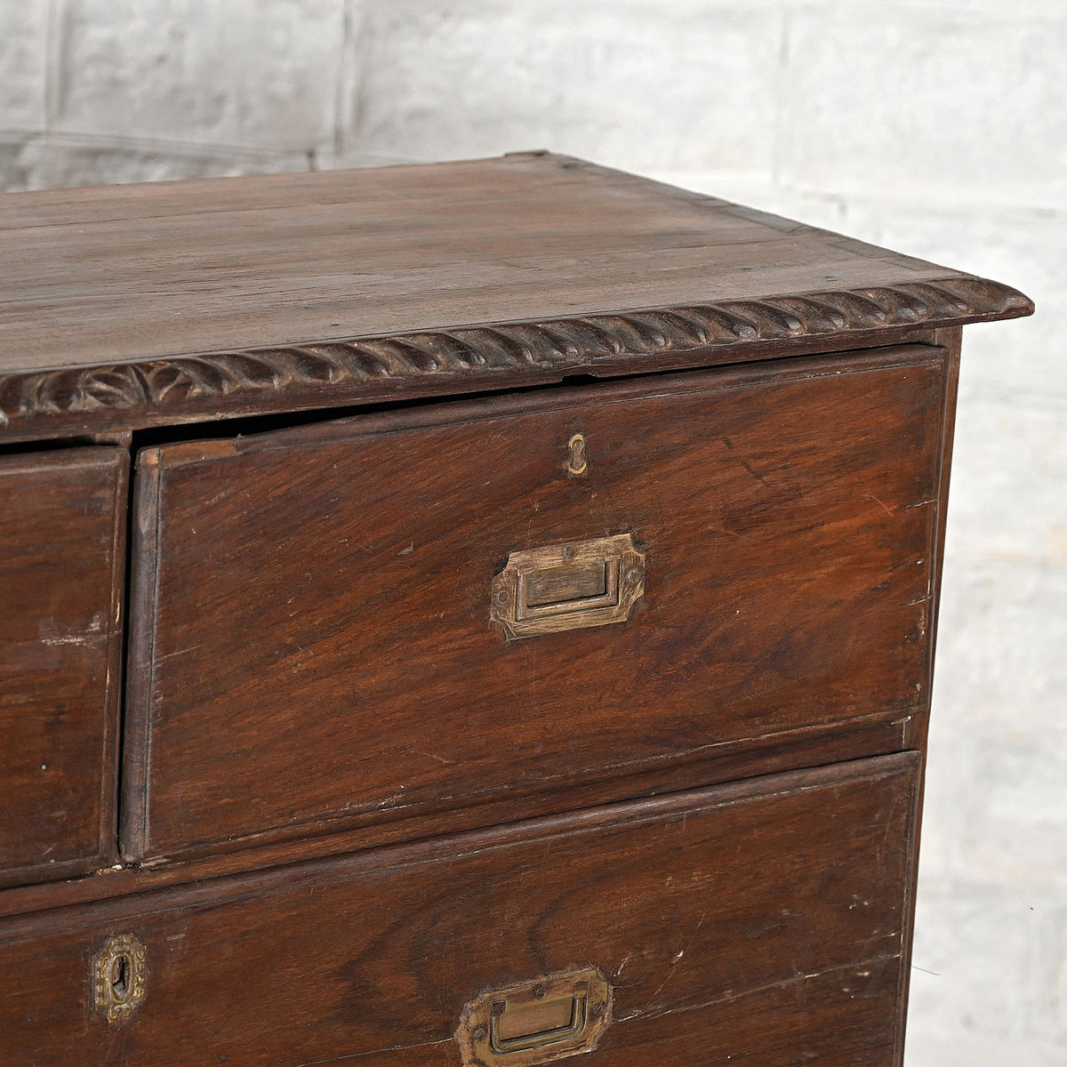 Close-up of the John Robshaw Teak Bureau 4, crafted from solid teak with ornate carved edges and brass label holders on its drawers, set against a light-colored brick wall.