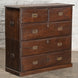 The John Robshaw Teak Bureau 4 is a vintage-inspired wooden chest of drawers crafted from solid teak in the style of classic campaign furniture. It features elegant brass handles, with two small top drawers and three spacious lower drawers, and is pictured against a gray brick wall.