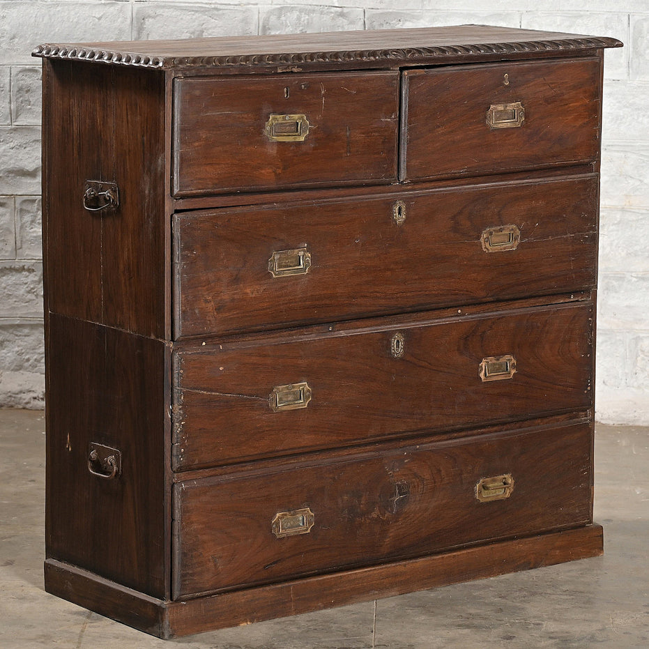 The John Robshaw Teak Bureau 4 is a vintage-inspired wooden chest of drawers crafted from solid teak in the style of classic campaign furniture. It features elegant brass handles, with two small top drawers and three spacious lower drawers, and is pictured against a gray brick wall.