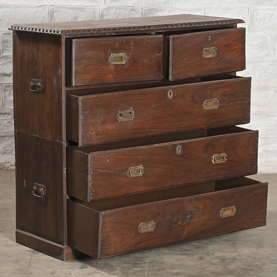 The John Robshaw Teak Bureau 4 is a vintage solid teak wooden chest of drawers, featuring five partially open drawers with brass handles and a decorative edge, set against a textured gray wall.
