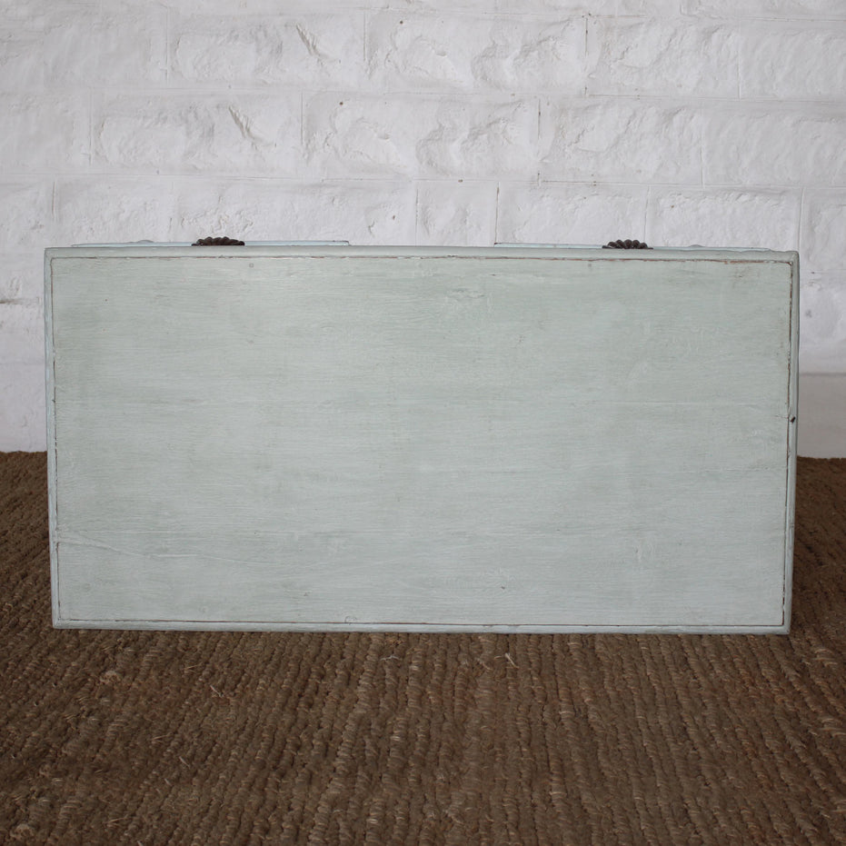 A rectangular, pale wooden Pravah Teak Desk by John Robshaw, with a flat lid closed, sits on a woven brown rug in front of a white brick wall, evoking the timeless charm of vintage design.