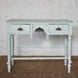 The Pravah Teak Desk by John Robshaw features a light blue vintage finish, two drawers with ornate brass cup pulls, and stands on four turned legs. It is shown placed on a woven rug against a white brick wall.