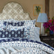 A neatly made bed featuring John Robshaw’s Diba Sand Performance Fabric bedding in blue and white patterns, complemented by a floral headboard, a bedside table with a lamp and flowers, and a wall painting of two birds—believed to bring good fortune and ward off the evil eye.