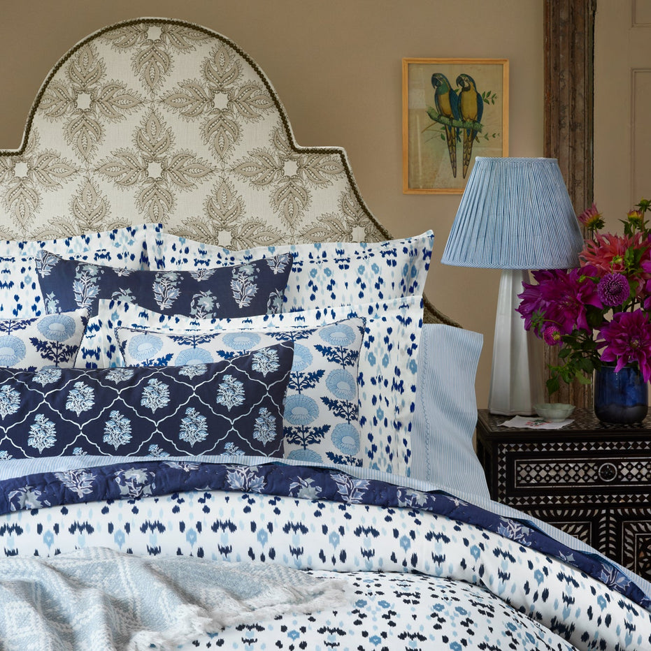 A neatly made bed featuring John Robshaw’s Diba Sand Performance Fabric bedding in blue and white patterns, complemented by a floral headboard, a bedside table with a lamp and flowers, and a wall painting of two birds—believed to bring good fortune and ward off the evil eye.