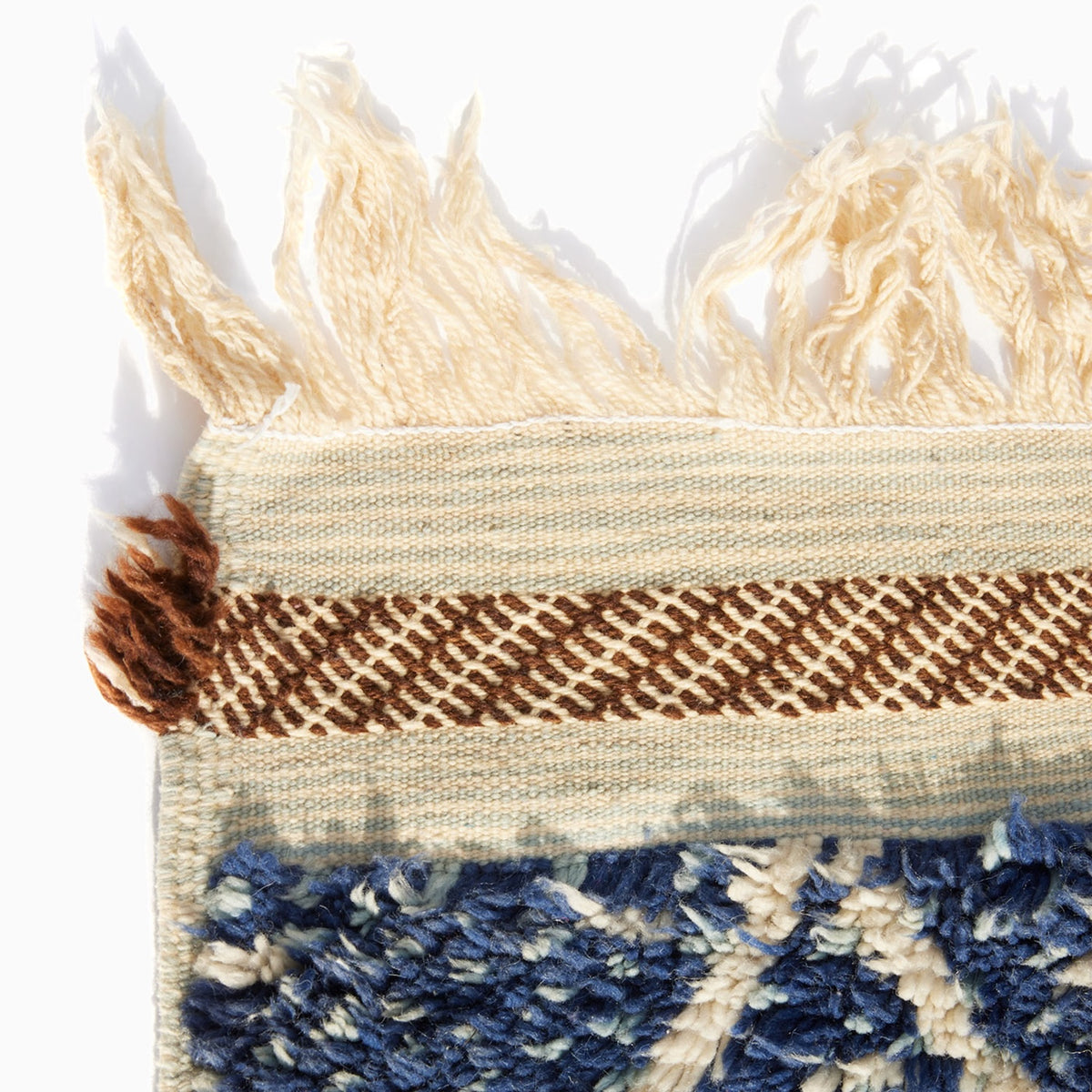 Corner of a Blue Village Wool Rug featuring a blue and cream patterned pile, a brown and cream geometric border, and long fringe at the top edge.